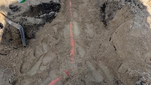 Gravested med en rød linje i sandet og en skovl ved siden af, der viser et byggeprojekt.