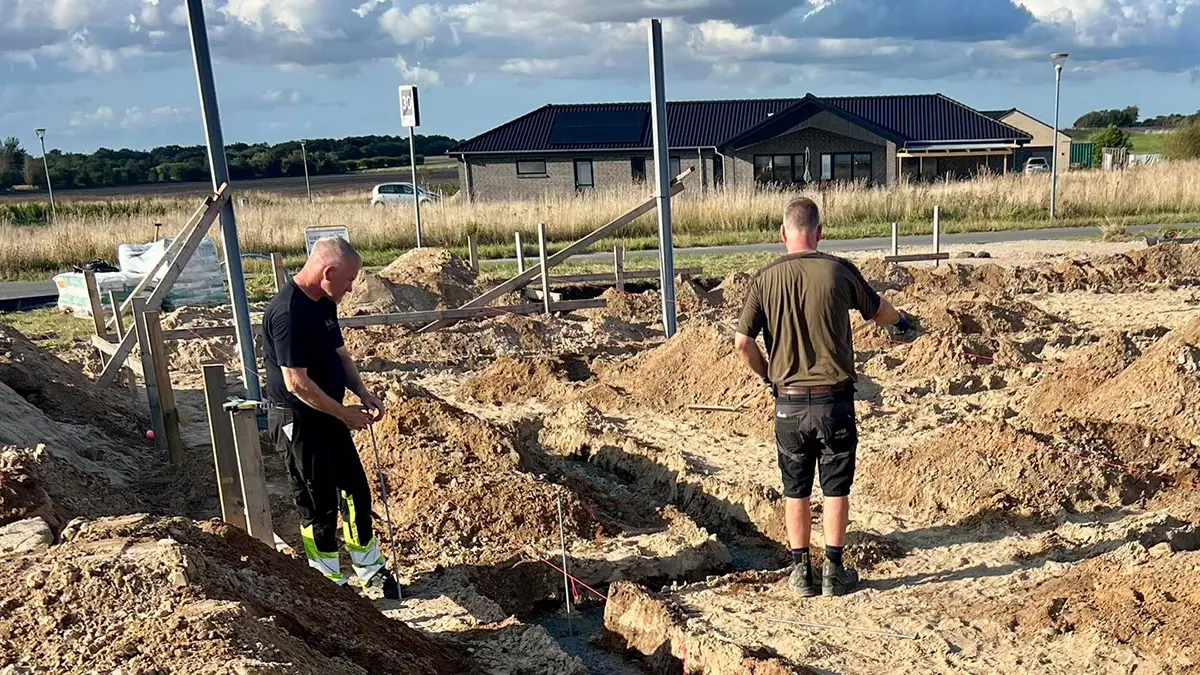 To mænd arbejder på en byggeplads med grus og fundament, mens en bolig ses i baggrunden.