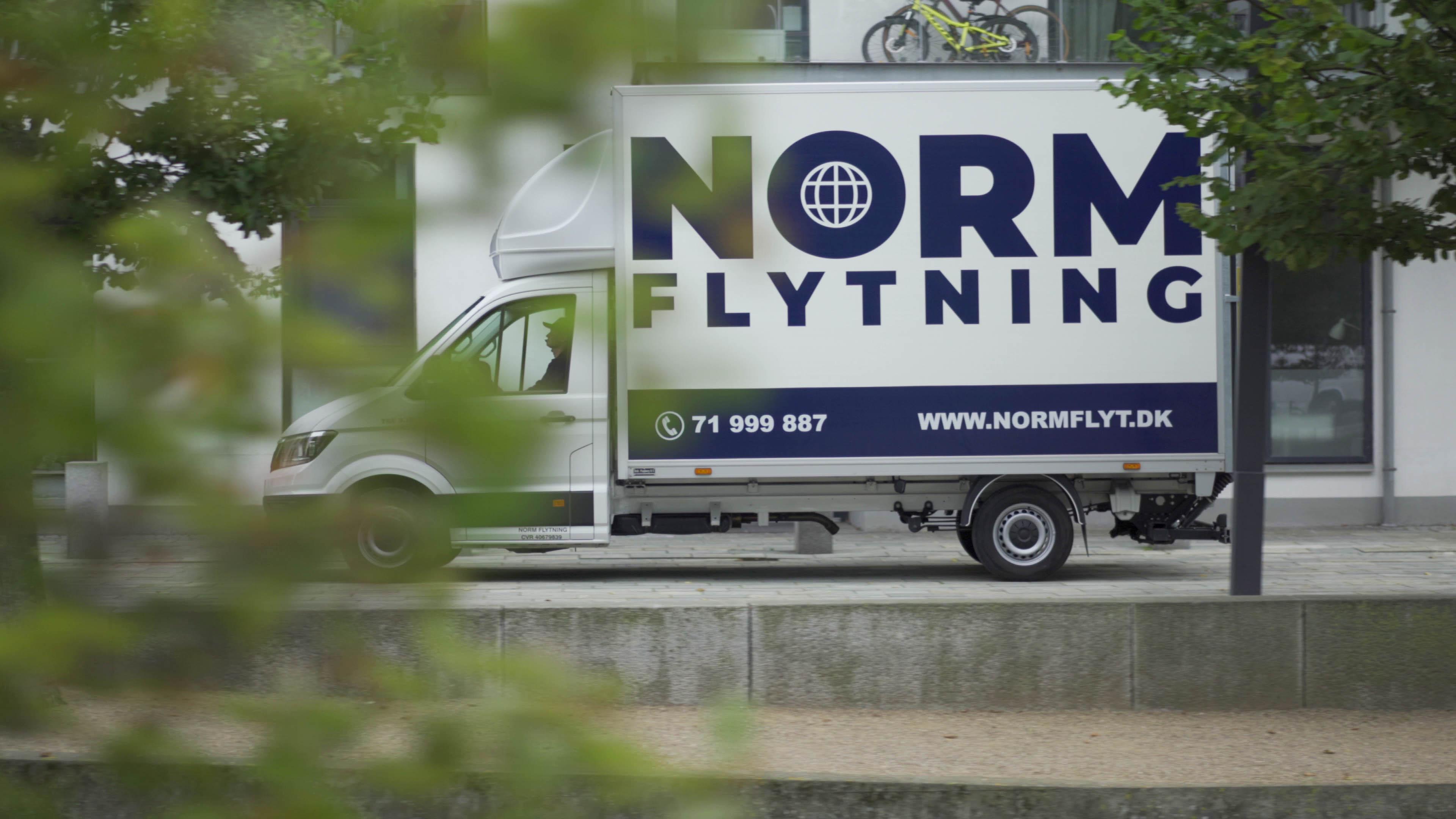 En flyttebil med teksten "NORM FLYTNING" og webadressen "www.NORMFLYT.dk" parkeret ved en bygning.