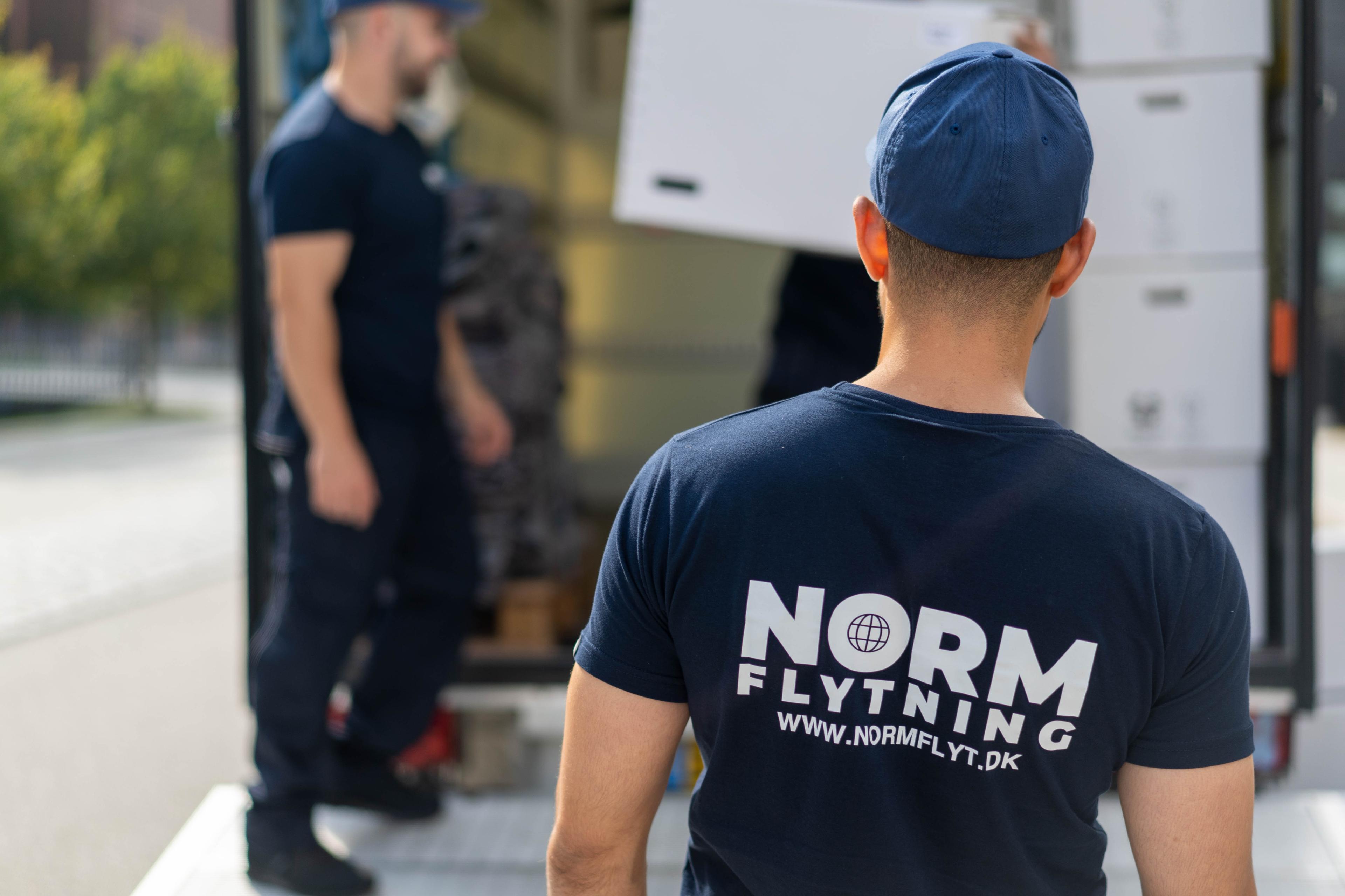 To mænd i arbejdstøj, der flytter kasser fra en lastbil, med fokus på en mand med ryggen til.