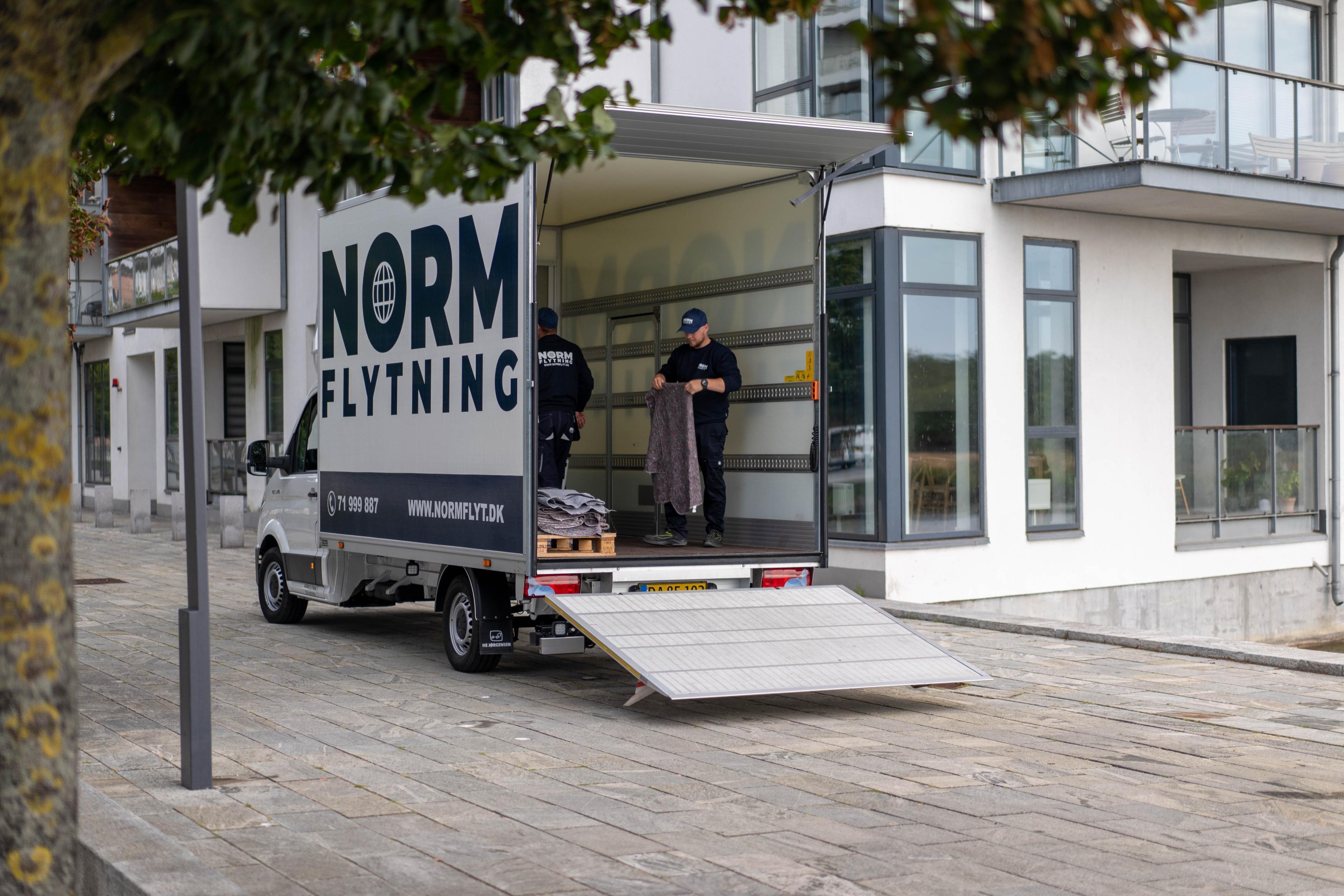 En flyttebil fra Norm Flytning står parkeret ved en moderne bygning, mens to personer laster kasser ind.