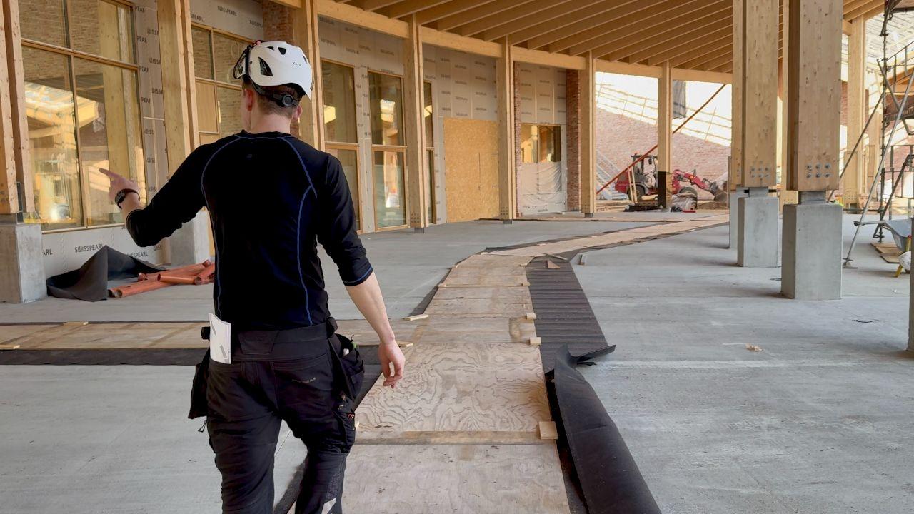 En arbejder iført hjelm går på en byggeplads med en lang rulle af sort materiale på gulvet.