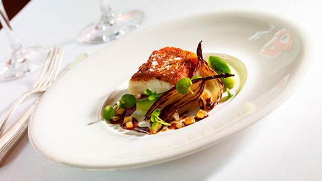 En elegant tallerken med stegt fisk, grøntsager og nødder, præsenteret smukt på en hvid dug.