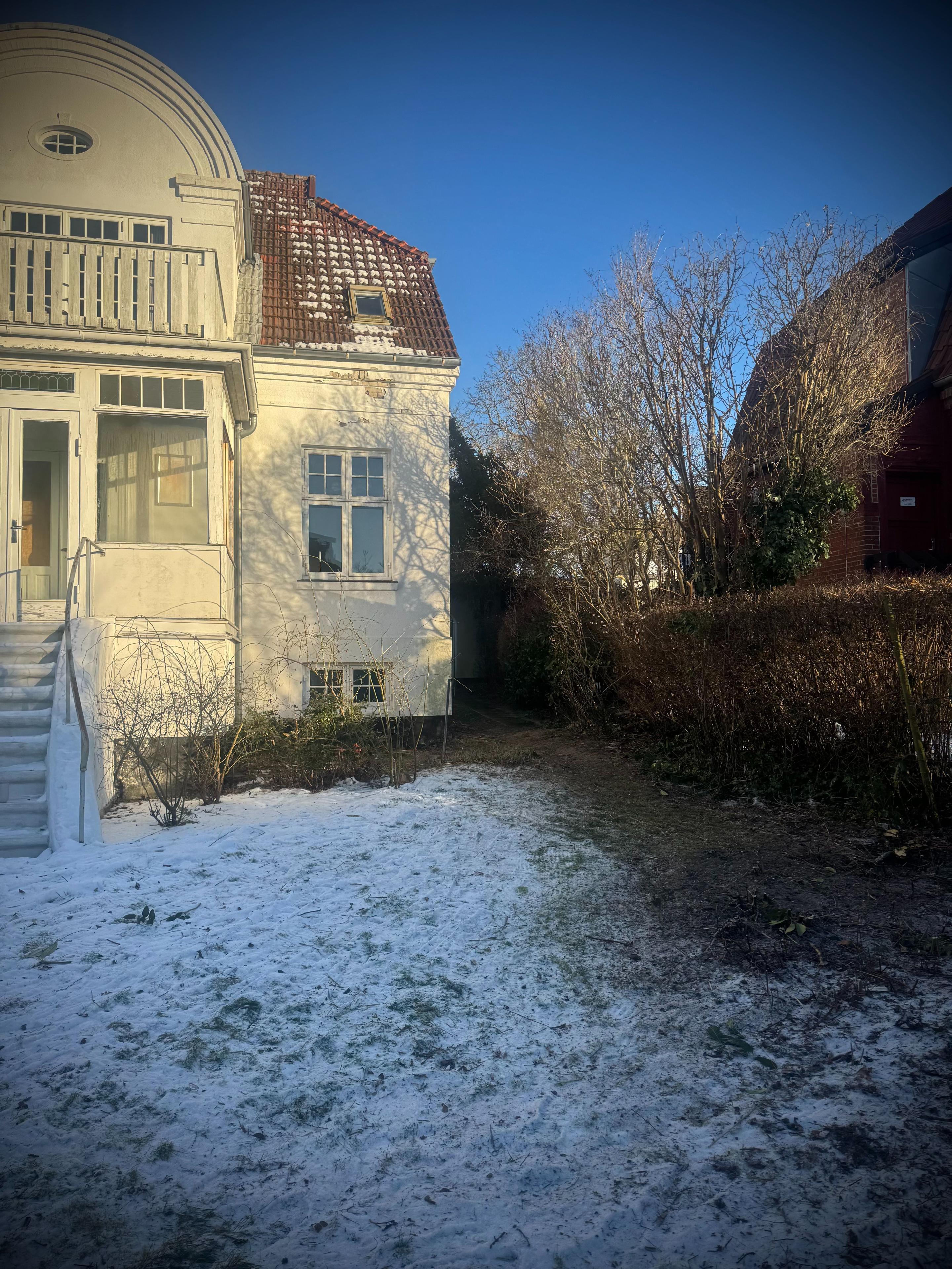 En hvid villa med en sneklædt have og buske i baggrunden under en klar blå himmel.