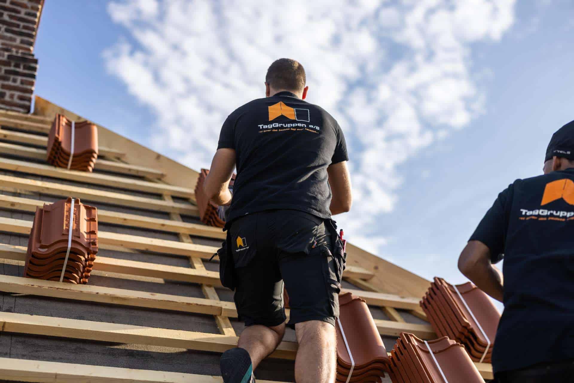 To håndværkere arbejder på et tag med teglsten under en blå himmel.