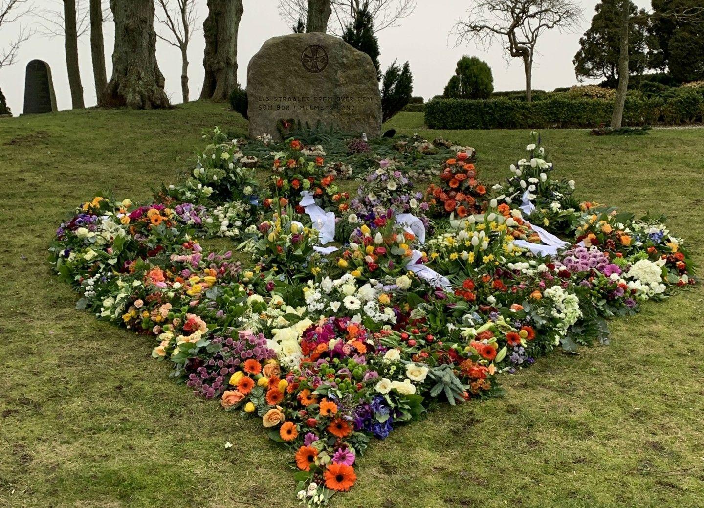 En farverig blomsterkrans i hjerteform på en gravplads med en stor sten i baggrunden.