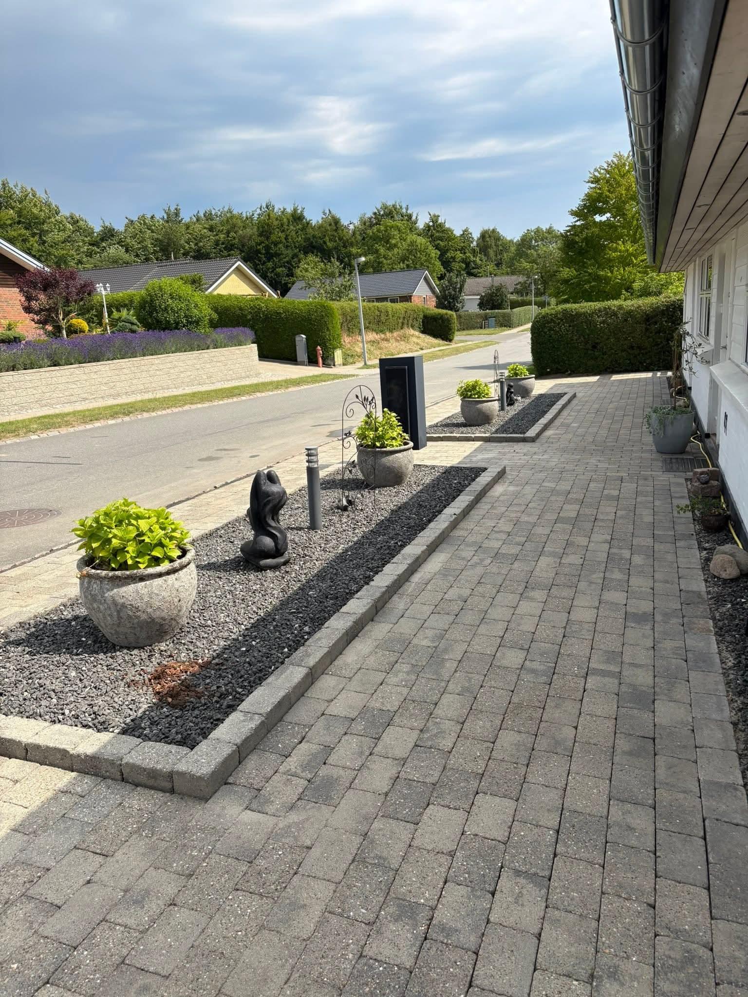 En indkørsel med grå fliser, planter i krukker og en sort kattefigur ved siden af en postkasse.