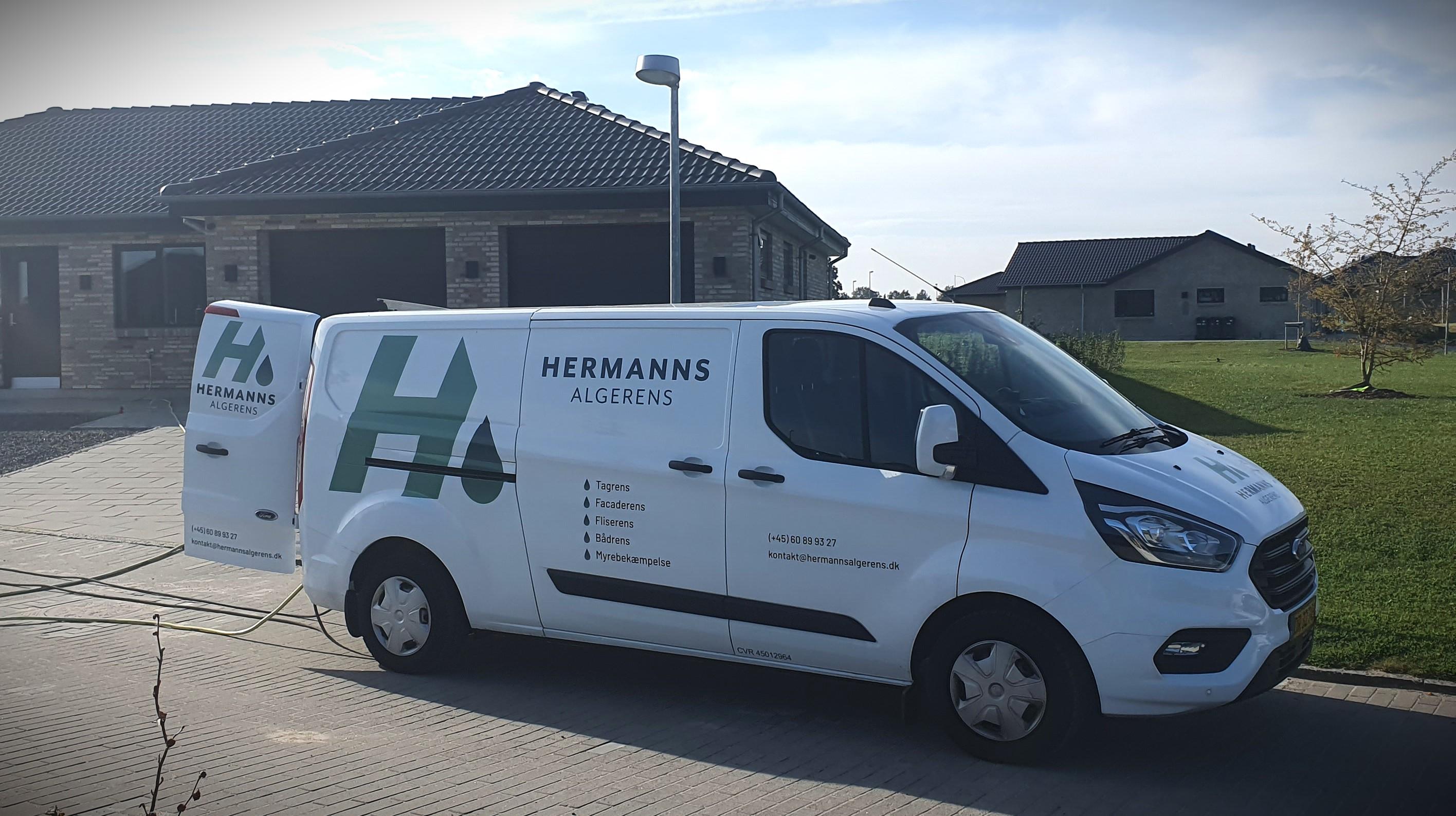 En hvid varevogn med logoet "Hermann's" parkeret foran et hus på en græsplæne.