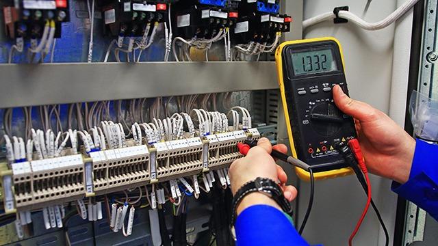 En person måler elektrisk spænding med et multimeter i et kontrolpanel med ledninger.