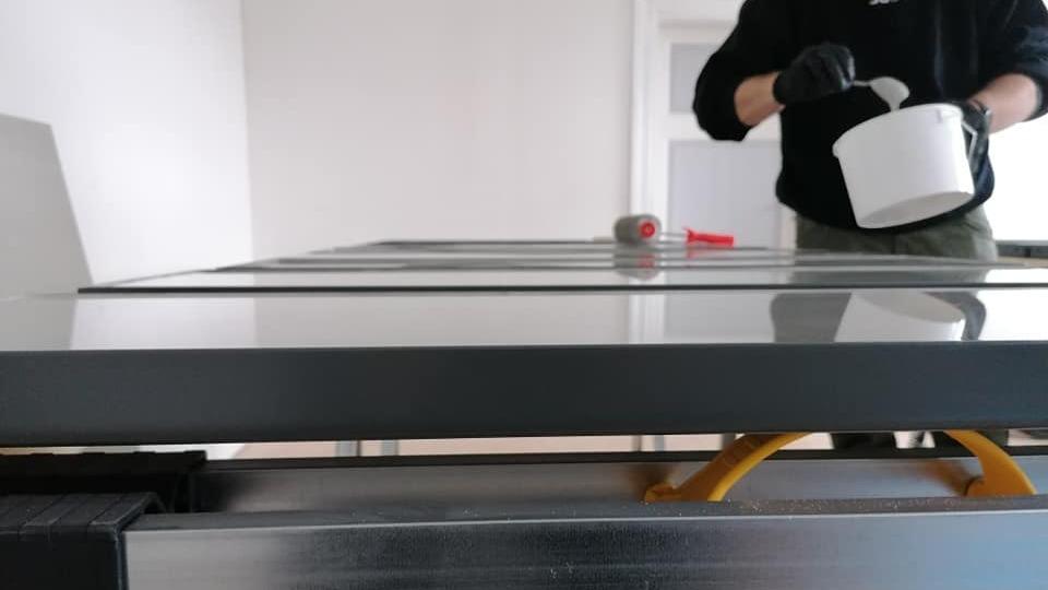 En person iført handsker hælder maling fra en spand på et bord i et lyst rum.