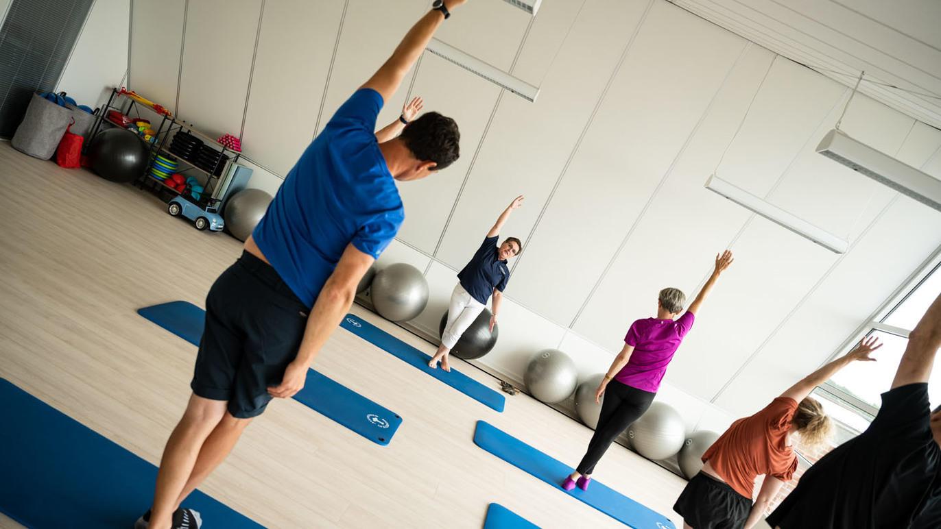 Gruppe af mennesker, der laver strækøvelser i et lyst træningslokale med træningsmåtter og fitnessbolde.