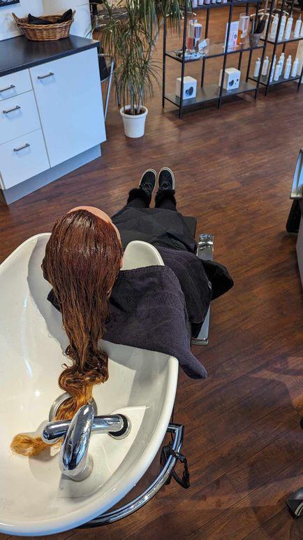 En person med langt, brunt hår ligger i en frisørsalon ved en vask, mens håret bliver vasket.