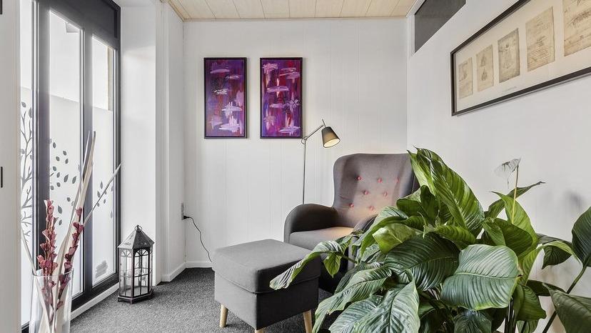 En hyggelig stue med en grå lænestol, en fodskammel, planter og farverige kunstværker på væggen.