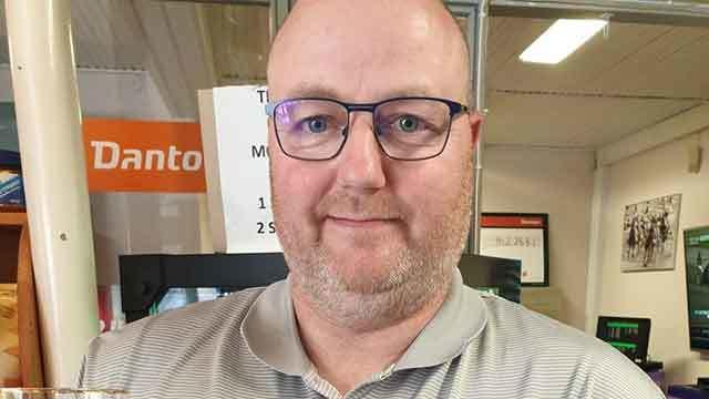 En mand med briller og skæg står foran en computer i et kontormiljø.