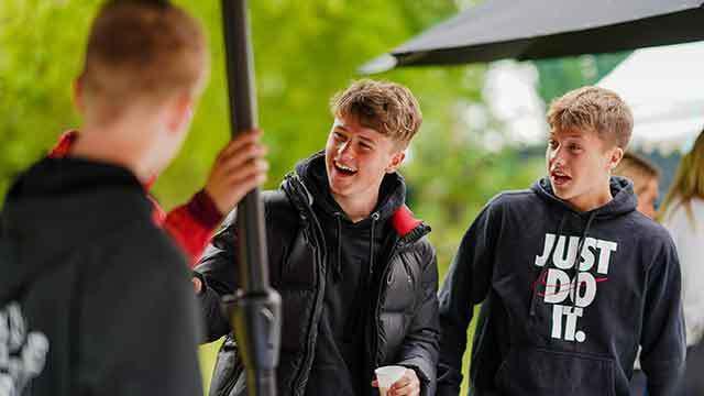 Tre unge mænd griner og snakker under en parasol, iført afslappet tøj og hættetrøjer.