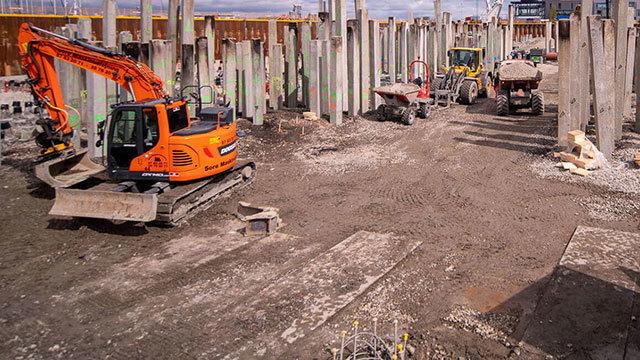 Byggeplads med gravemaskiner og lastbiler, der arbejder på fundamentet af en bygning under konstruktion.