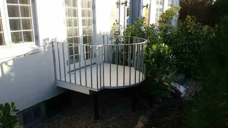 En rund terrasse med gelænder ved en hvid bygning omgivet af grønne planter.