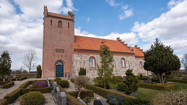 En kirke med rød tag og murstensvægge omgivet af gravsteder og grønne buske under en blå himmel.