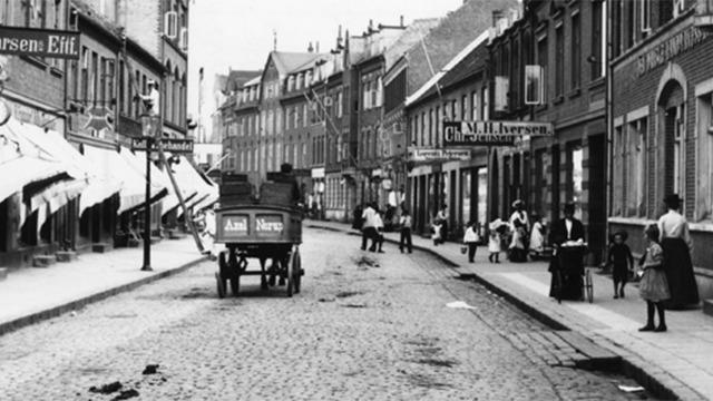 En historisk gade med brosten, hestevogn og mennesker, der går forbi butikker og caféer.