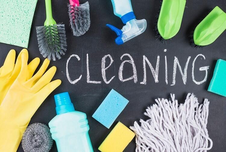 Rengøringsartikler som svampe, børster, handsker og sprayflaske med ordet "CLEANING" skrevet på en tavle.