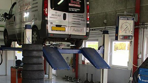 En bil på en lift i et værksted med dæk og bremsetestudstyr i baggrunden.