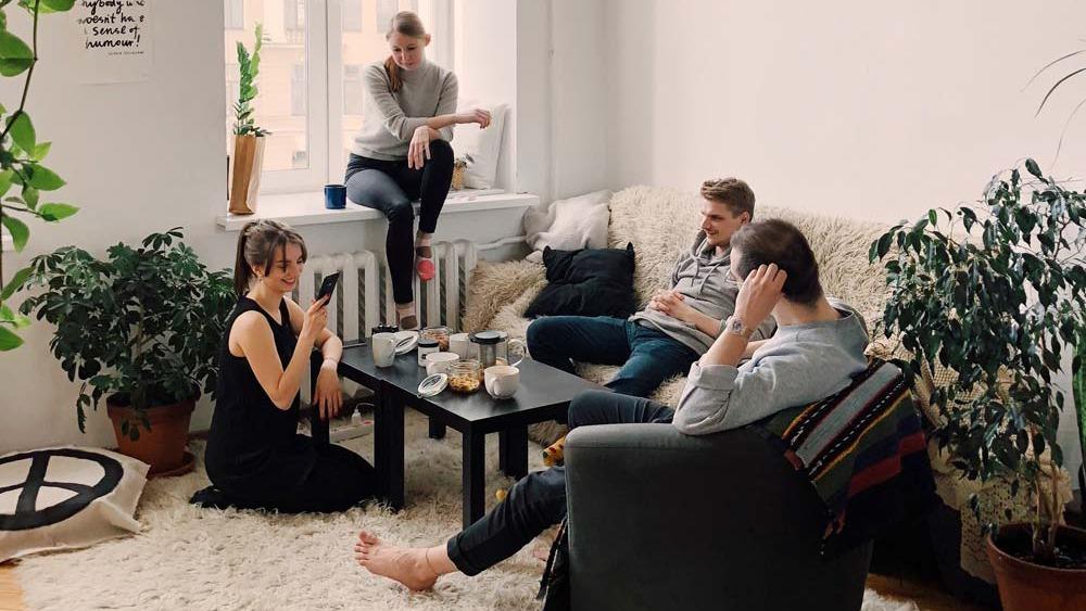 Fire unge mennesker sidder i en stue med planter, drikker kaffe og snakker sammen på en hyggelig sofa.
