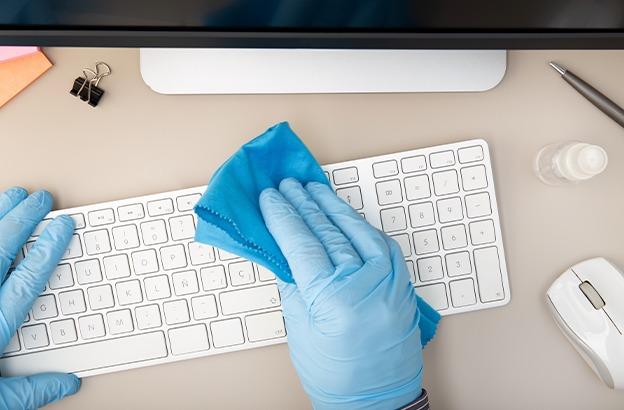 Hænder iført handsker rengør et tastatur med en blå klud og desinfektionsmiddel på et skrivebord.