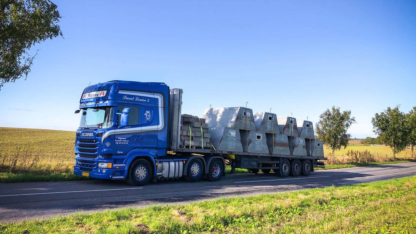 En blå lastbil transporterer betonblokke på en landevej med marker i baggrunden.