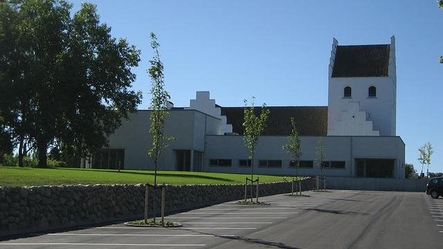 Moderne kirke med hvid facade og klokketårn, omgivet af grønne træer og parkeringsplads.