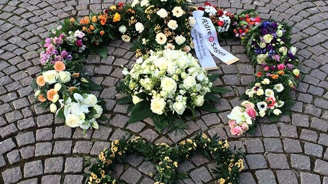 Blomsterkranse i form af et fredssymbol lavet af farverige blomster på en brostensbelagt overflade.