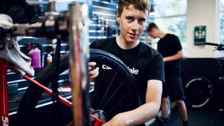 En ung mand reparerer en cykel i en cykelbutik, mens en anden person arbejder i baggrunden.