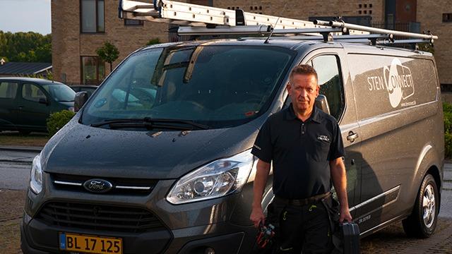 En mand står ved siden af en grå varevogn med tagræling, klar til arbejde i et boligområde.