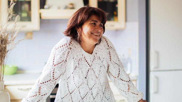 Kvinde med brunt hår iført en hvid strikket sweater, smilende i et lyst køkken.