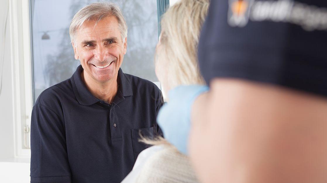 En smilende mand i en sort polo t-shirt sidder overfor en kvinde i et lyst rum.