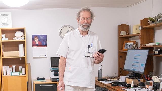En ældre mand i hvidt lægetøj står i et kontor med en telefon i hånden og ser mod kameraet.