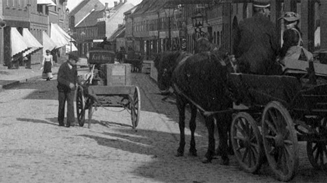 Gammel gade med hestevogn og mennesker, der arbejder med en vogn på brostenene.