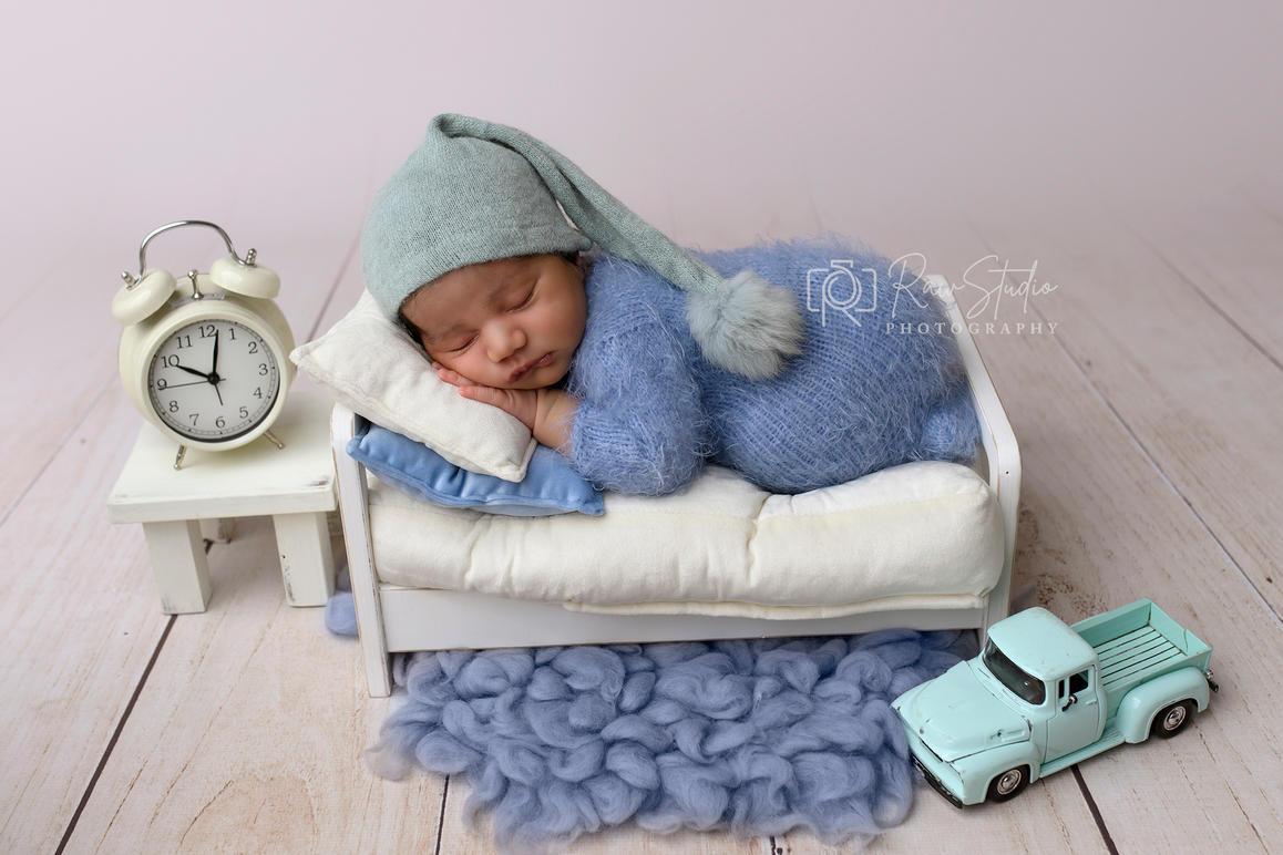 Nyfødt baby sover fredeligt i en lille seng med en blå hue, ved siden af en vækkeur og en legetøjsbil.