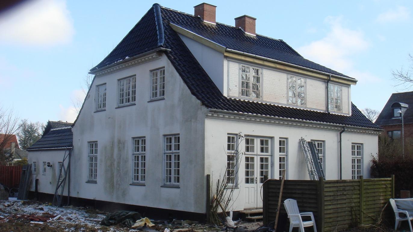 Et hvidt hus med sort tag og mange vinduer, omgivet af en have med byggeaffald og en stol.
