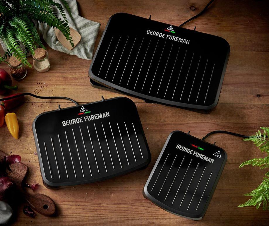 Tre George Foreman grillplader i forskellige størrelser på et træbord med grøntsager i baggrunden.