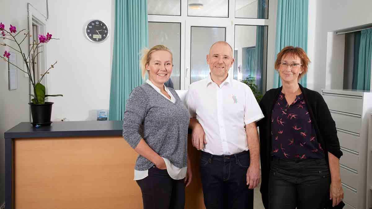 Tre personer står ved en reception med en plante i baggrunden og et ur på væggen.
