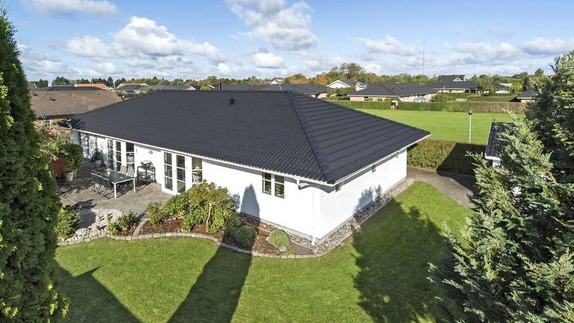 Et moderne hus med sort tag, omgivet af grøn græsplæne og buske, set fra luften.