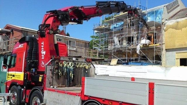 En rød lastbil med kran arbejder på en byggeplads med stilladser og materialer.