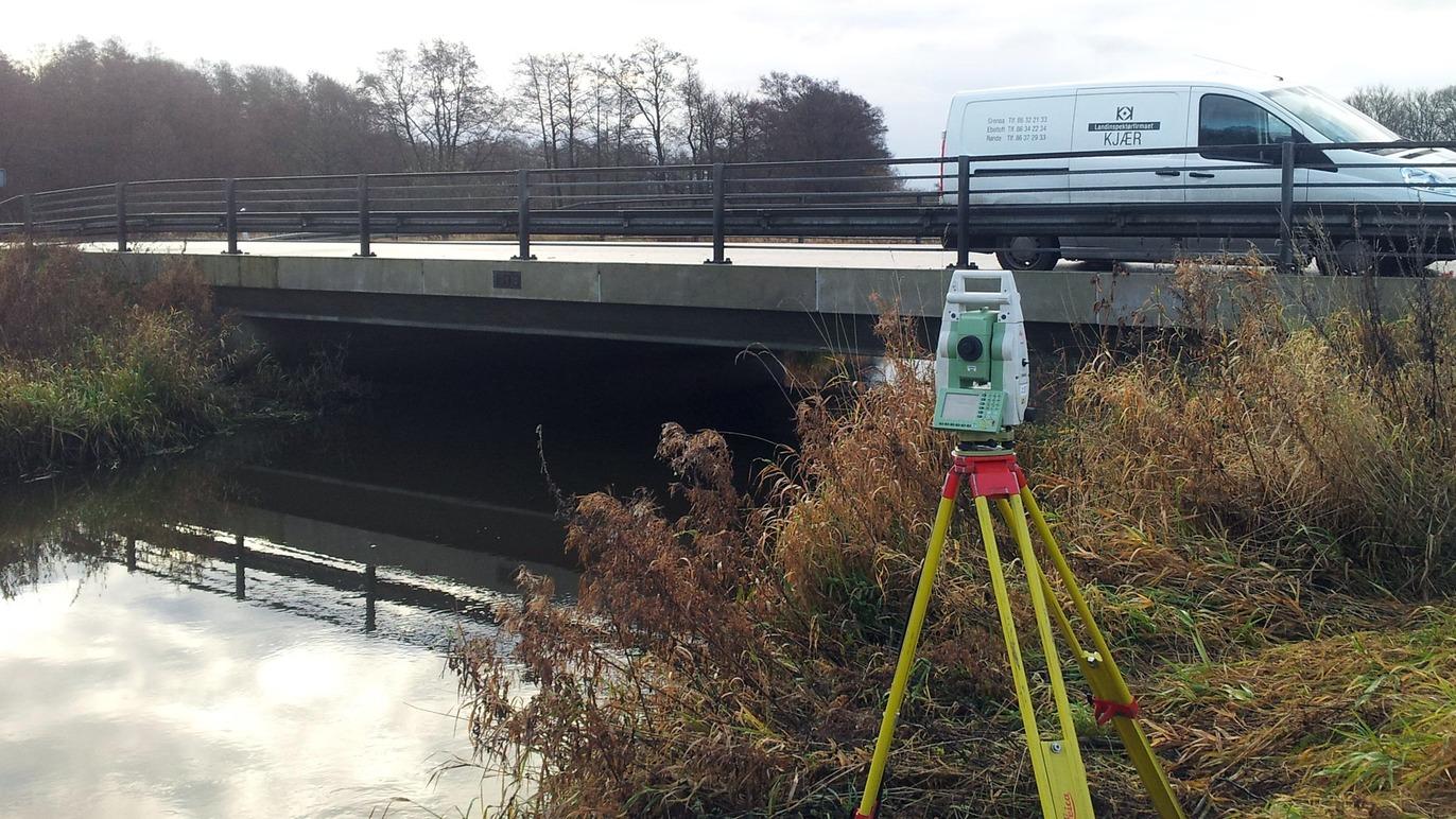 En totalstation står på en stativ ved en bro over en stille vandløb med en varevogn i baggrunden.