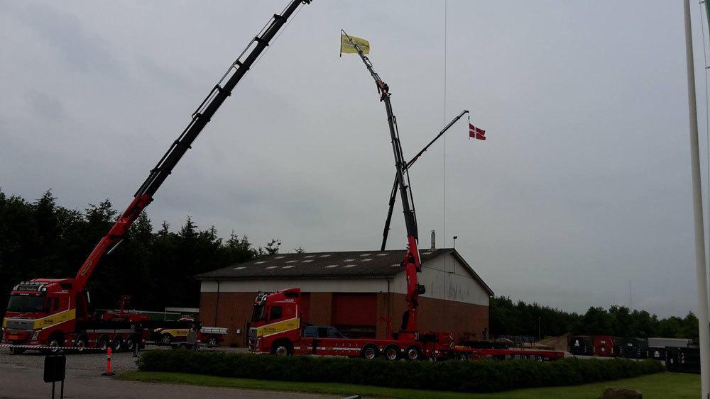 To store med kraner, der løfter flag over en bygning, med lastbiler parkeret i forgrunden.