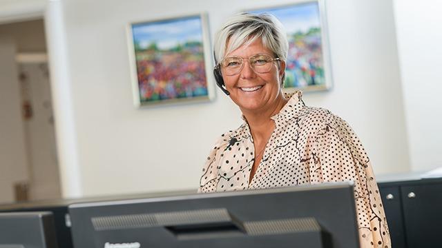 En smilende kvinde med headset står ved en computer i et kontormiljø med malerier i baggrunden.