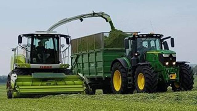 En grøn høstmaskine og en traktor, der transporterer græs fra marken til en vogn.