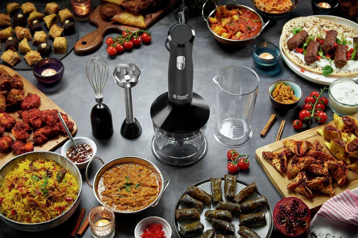 Et bord dækket med forskellige retter, herunder ris, kød, grøntsager og snacks, samt en stavblender og køkkenredskaber.