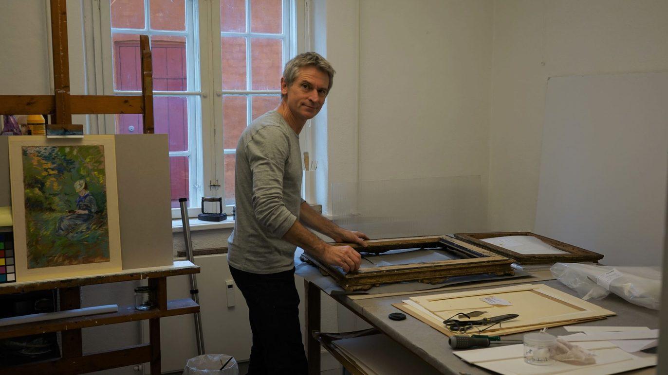 En kunstner arbejder med rammer i sit atelier, omgivet af malerier og kunstmaterialer.