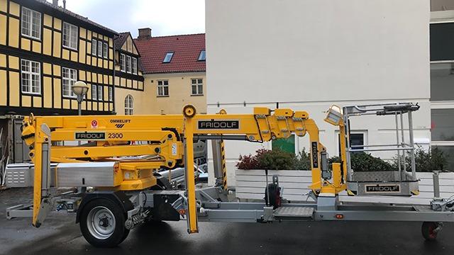 En gul lift på en trailer parkeret ved en bygning med bindingsværk.