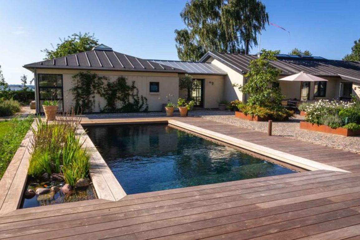 Moderne hus med swimmingpool og træterrasse omgivet af planter og blomster i en solrig have.